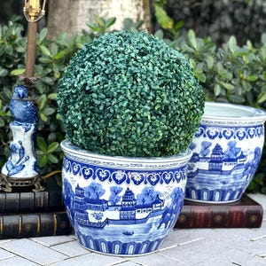 May include: A blue and white ceramic planter with a detailed landscape design, holding a spherical green plant. The planter is placed next to another similar planter and a decorative lamp.