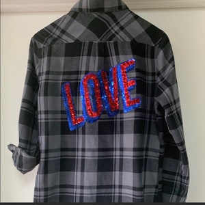 Może przedstawiać: Koszula flanelowa w szaro-czarną kratę z naszywką "LOVE" z czerwonych i niebieskich cekinów na plecach.