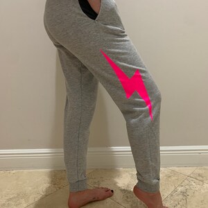 Puede incluir: Pantalones de ch&aacute;ndal grises con un gr&aacute;fico de rayo rosa en la pierna derecha. Los pantalones de ch&aacute;ndal tienen una cintura negra y un cord&oacute;n negro.