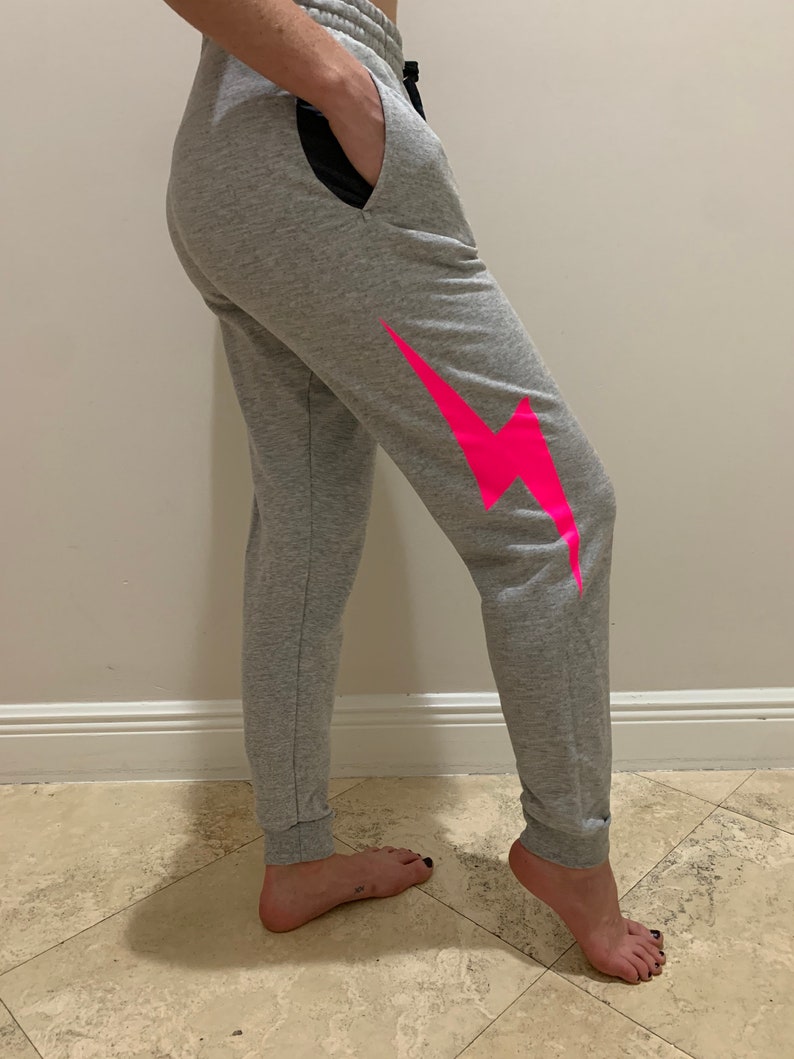 Puede incluir: Pantalones de ch&aacute;ndal grises con un gr&aacute;fico de rayo rosa en la pierna derecha. Los pantalones de ch&aacute;ndal tienen una cintura negra y un cord&oacute;n negro.