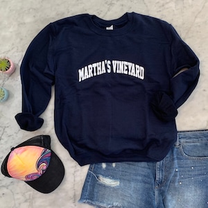 May include: A navy blue crewneck sweatshirt with the words "MARTHA'S VINEYARD" in white arched lettering. The sweatshirt is paired with denim shorts and a colorful trucker hat. The items are arranged on a marble surface.