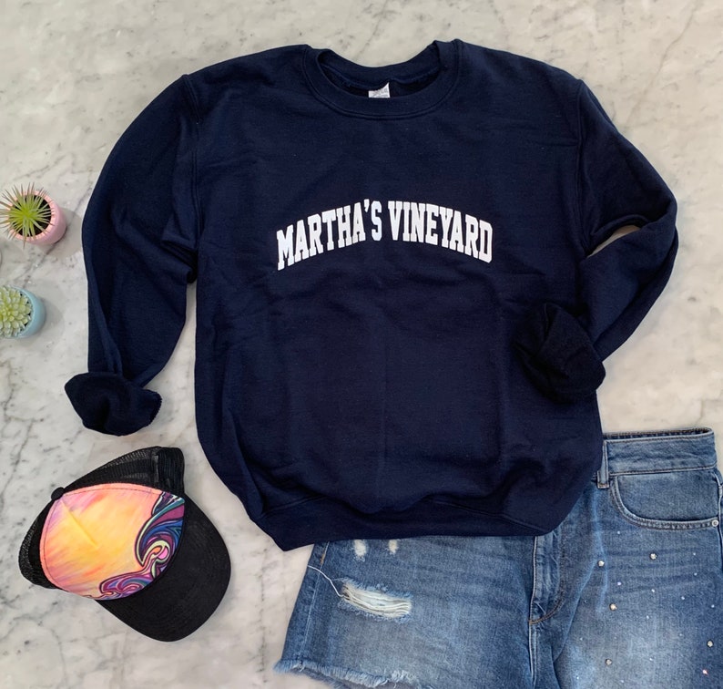 May include: A navy blue crewneck sweatshirt with the words "MARTHA'S VINEYARD" in white arched lettering. The sweatshirt is paired with denim shorts and a colorful trucker hat. The items are arranged on a marble surface.