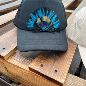 Puede incluir: Gorra de camionero negra de malla con un dise&ntilde;o de plumas azules, doradas y negras en la parte delantera.