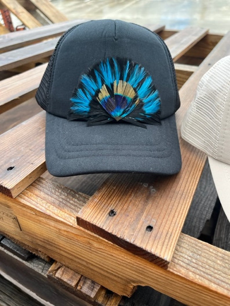 Puede incluir: Gorra de camionero negra de malla con un dise&ntilde;o de plumas azules, doradas y negras en la parte delantera.