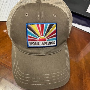 May include: A brown and beige mesh baseball cap with a colourful sunrise and the text "Hola Amigos" embroidered on the front.