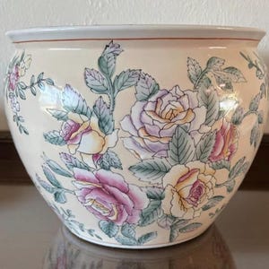 May include: A cream-colored ceramic planter with a floral rose design. The planter features pink, yellow, and purple roses with green leaves. The rim has a thin red stripe.