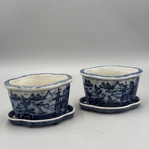 May include: Two oval ceramic planters with matching saucers, featuring a blue and white transferware landscape pattern depicting pagodas, trees, and mountains. The off-white interior surfaces show a fine crackle glaze finish throughout.