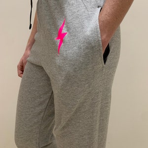Puede incluir: Pantalones de ch&aacute;ndal grises con un gr&aacute;fico de rayo rosa. Los pantalones de ch&aacute;ndal tienen cintura con cord&oacute;n y bolsillos laterales.