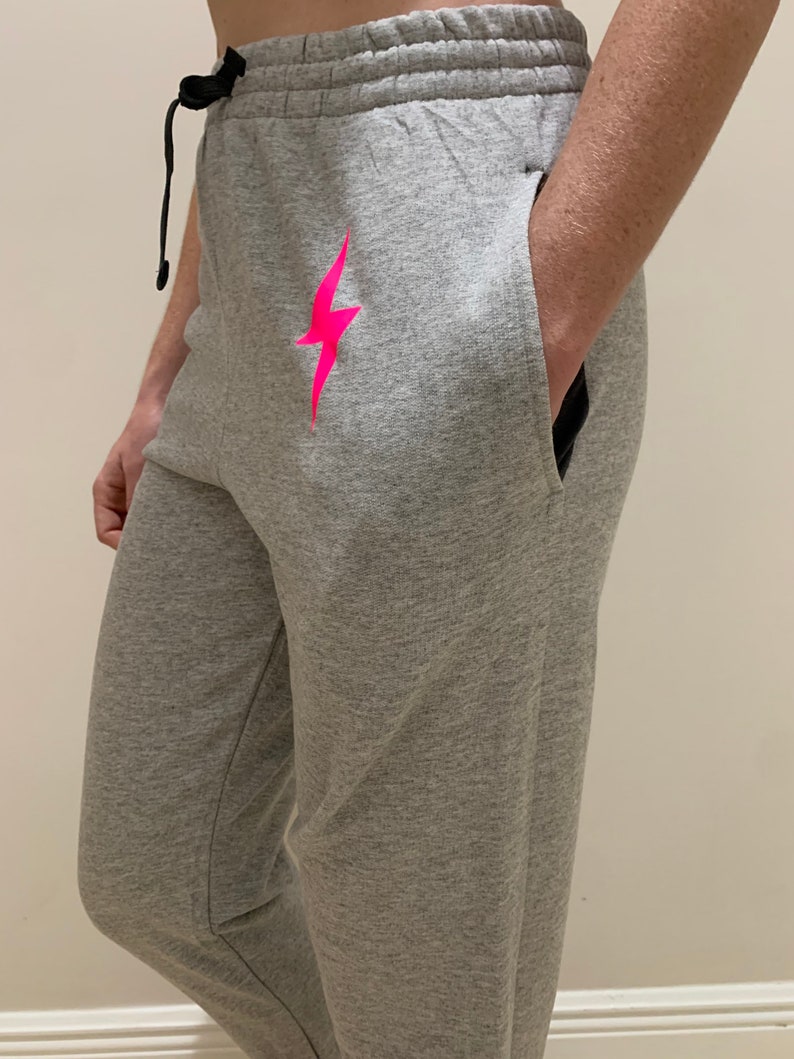 Puede incluir: Pantalones de ch&aacute;ndal grises con un gr&aacute;fico de rayo rosa. Los pantalones de ch&aacute;ndal tienen cintura con cord&oacute;n y bolsillos laterales.