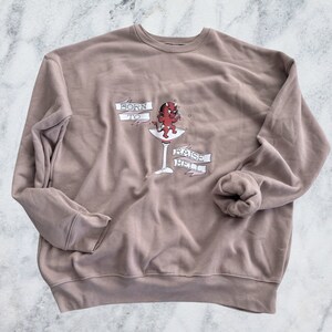 May include: A taupe-colored sweatshirt with a graphic of a red devil in a martini glass. The text "BORN TO RAISE HELL" is displayed in banners around the glass. The sweatshirt has a crew neck and long sleeves.