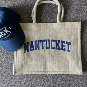 May include: A beige jute tote bag with navy blue rope handles and the word "NANTUCKET" printed in navy blue. A navy blue baseball cap with a white oval logo is in the background.