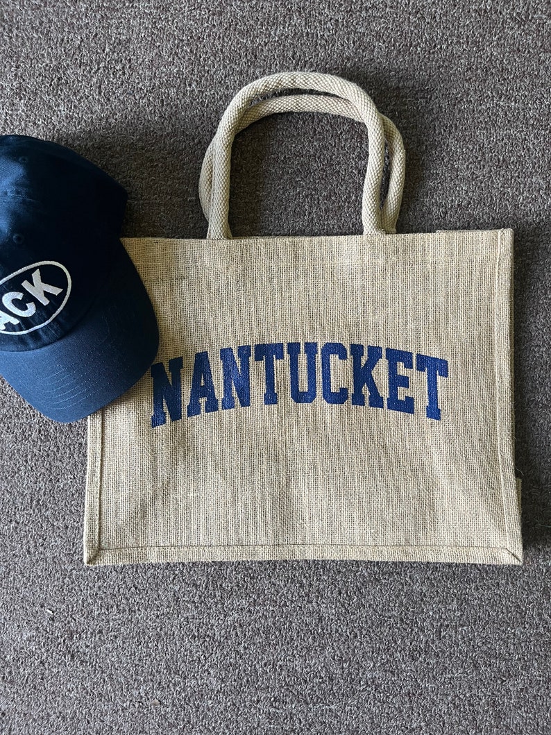 May include: A beige jute tote bag with navy blue rope handles and the word "NANTUCKET" printed in navy blue. A navy blue baseball cap with a white oval logo is in the background.
