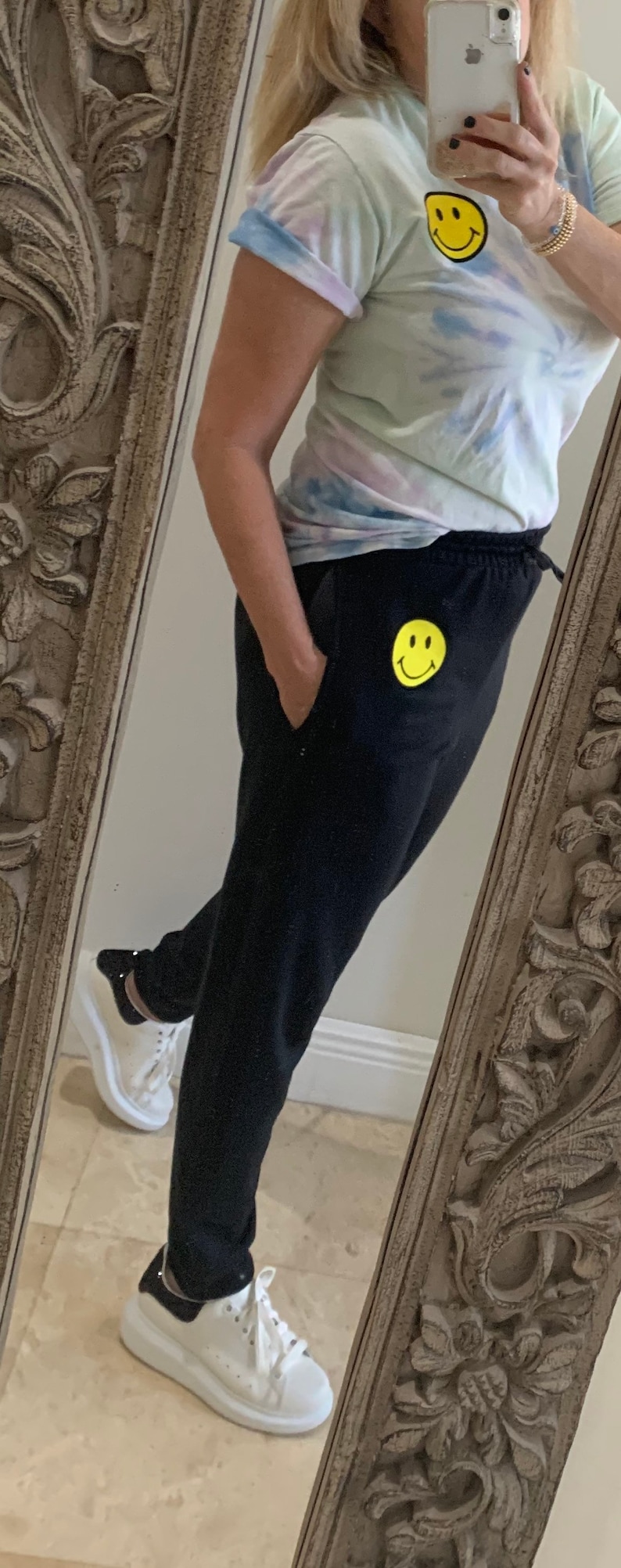 Puede incluir: Una mujer con pantalones de ch&aacute;ndal negros con un parche de cara sonriente amarilla en la pierna izquierda. Tambi&eacute;n lleva una camiseta de color azul claro con estampado tie-dye con un parche de cara sonriente amarilla en el lado derecho. Lleva zapatillas blancas con ribetes negros.