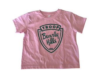 May include: Pink short-sleeved t-shirt with a green shield graphic that says "TROOP Beverly Hills" in a retro font.