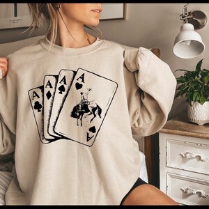 May include: A beige sweatshirt with a graphic of four playing cards, each showing the Ace and a different suit. The Ace of Spades card features a cowboy riding a horse.