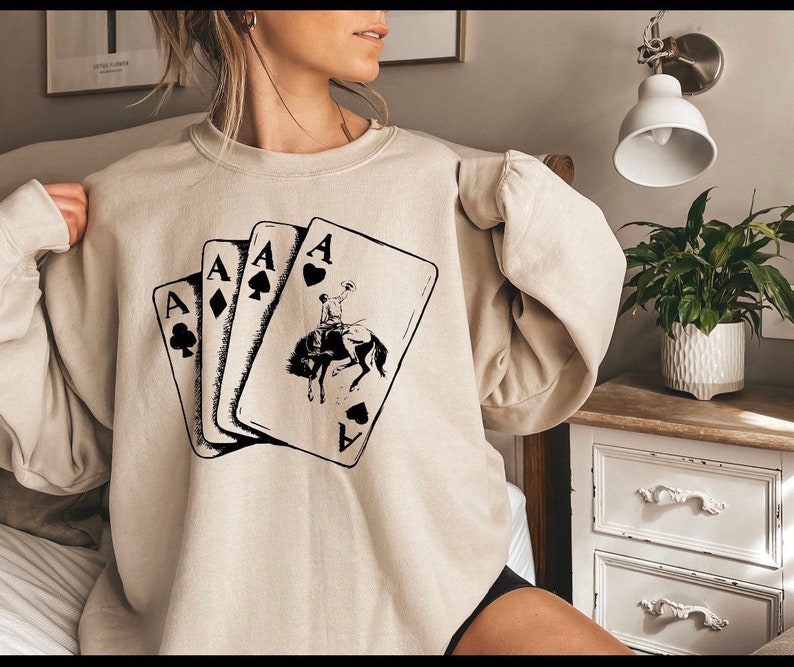 May include: A beige sweatshirt with a graphic of four playing cards, each showing the Ace and a different suit. The Ace of Spades card features a cowboy riding a horse.
