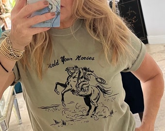May include: A sage green t-shirt with a black graphic of a woman riding a horse and the text "Hold Your Horses".