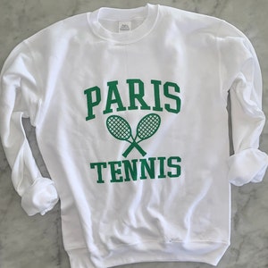 May include: White sweatshirt with green text and tennis rackets. The word "PARIS" is arched above the crossed tennis rackets, with "TENNIS" below. The long sleeves are cuffed.