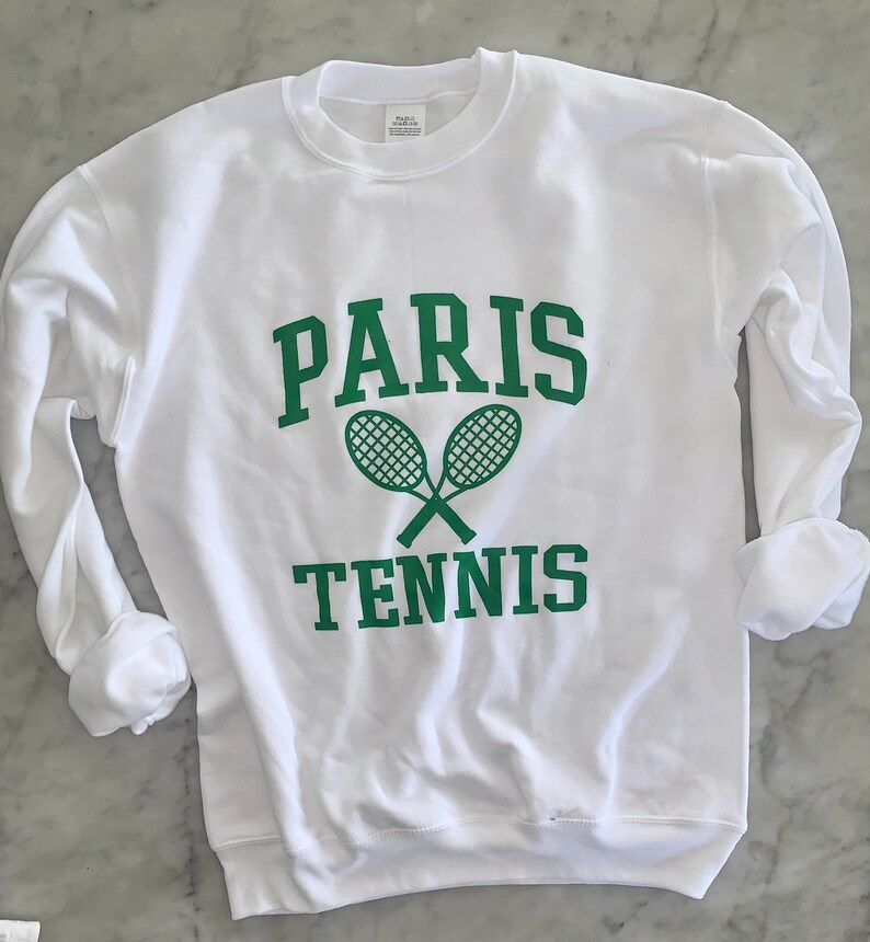May include: White sweatshirt with green text and tennis rackets. The word "PARIS" is arched above the crossed tennis rackets, with "TENNIS" below. The long sleeves are cuffed.