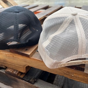 Puede incluir: Dos gorras de camionero de malla, una negra y otra beige. Ambas gorras tienen una malla en la parte posterior y un panel frontal s&oacute;lido.