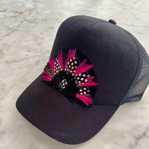 Puede incluir: Gorra de camionero negra con un adorno de plumas rosa y negro. La pluma tiene lunares blancos.