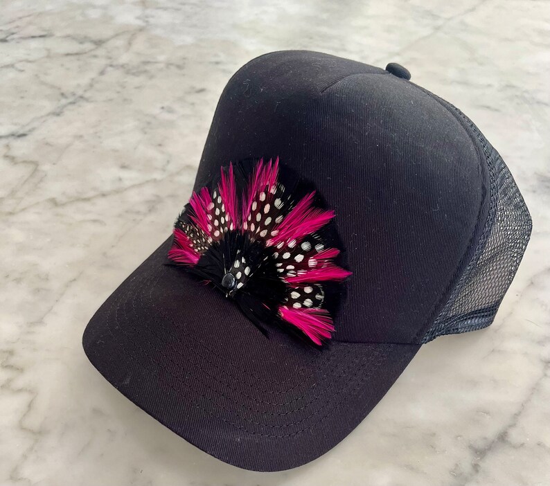 Puede incluir: Gorra de camionero negra con un adorno de plumas rosa y negro. La pluma tiene lunares blancos.