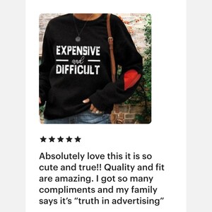 Peut inclure: Sweat-shirt noir &agrave; manches longues avec un patch en forme de c&oelig;ur rouge sur la manche gauche. Le texte "EXPENSIVE and DIFFICULT" est imprim&eacute; en blanc sur le devant.