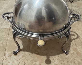 May include: A silver-colored, domed serving dish with ornate handles and a decorative base. The dish has a round, ivory-colored knob on the front. The base features curved legs. The background includes a metal rack and a tiled floor.