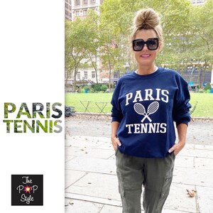 May include: A navy blue sweatshirt with the words "PARIS TENNIS" in white, along with crossed tennis rackets. The person is wearing sunglasses and olive green pants. The background includes a park with trees and buildings.