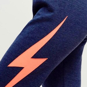 Puede incluir: Pantalones de ch&aacute;ndal azul marino con un gr&aacute;fico de rayo naranja brillante en la pierna izquierda.