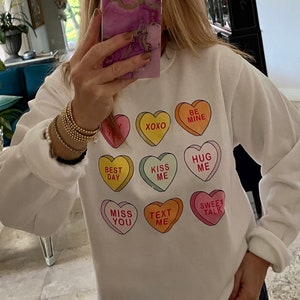 K&ouml;nnte beinhalten: Wei&szlig;es Sweatshirt mit bunten Bonbonherz-Grafiken. Jedes Herz hat eine andere Botschaft, darunter "XOXO", "Be Mine", "Kiss Me" und "Sweet Talk". Das Sweatshirt hat lange &Auml;rmel.
