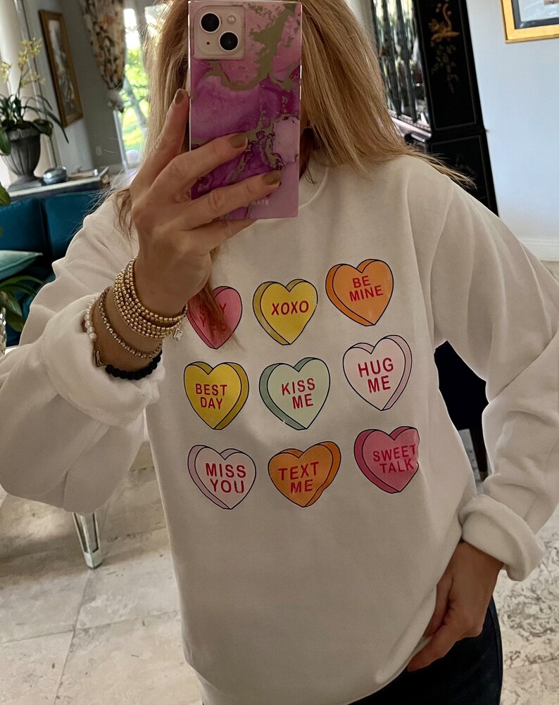 K&ouml;nnte beinhalten: Wei&szlig;es Sweatshirt mit bunten Bonbonherz-Grafiken. Jedes Herz hat eine andere Botschaft, darunter "XOXO", "Be Mine", "Kiss Me" und "Sweet Talk". Das Sweatshirt hat lange &Auml;rmel.