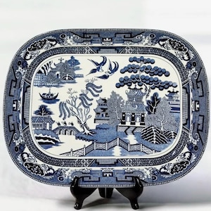 May include: An oval, blue and white ceramic serving platter with a Willow pattern design. The platter features a detailed landscape scene with pagodas, bridges, and birds. The border has a decorative geometric pattern. The platter is displayed on a black stand.