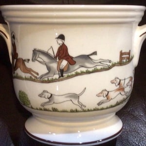 May include: Cream-colored ceramic planter with a hunting scene design. The planter features a wide, rounded body with two curved handles and a pedestal base. The illustration depicts a rider on horseback, hounds, and a fox.