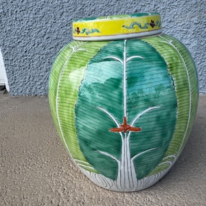 May include: A round, green and white ceramic jar with a lid. The jar features a central green leaf design with a white tree-like pattern and a red accent. The lid is yellow with floral accents. The jar is on a concrete surface.