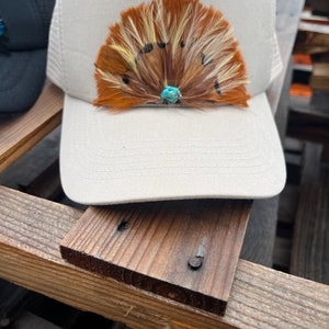 Puede incluir: Una gorra de camionero de malla blanca con un dise&ntilde;o de plumas marrones, blancas y amarillas. Una piedra turquesa se encuentra en el centro del dise&ntilde;o de plumas.