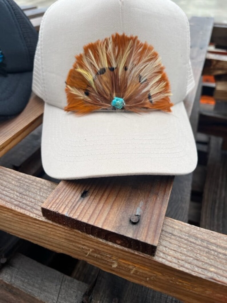 Puede incluir: Una gorra de camionero de malla blanca con un dise&ntilde;o de plumas marrones, blancas y amarillas. Una piedra turquesa se encuentra en el centro del dise&ntilde;o de plumas.