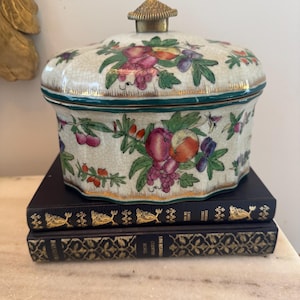 May include: A decorative ceramic lidded jar with a floral design featuring purple grapes, peaches, and green leaves. The jar has a gold rim and a brass knob on top. It sits on top of two dark blue books with gold lettering.