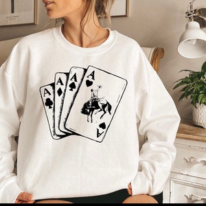 May include: White sweatshirt with a black graphic of four playing cards, each showing the Ace of a different suit. The Ace of Spades card features a cowboy riding a horse.