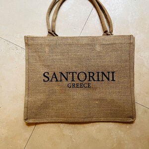 May include: A brown burlap tote bag with black handles and the text "SANTORINI GREECE" printed on the front.