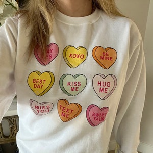 K&ouml;nnte beinhalten: Wei&szlig;es Sweatshirt mit bunten Bonbonherz-Grafiken mit Aufschriften wie "BE MINE" und "SWEET TALK". Die Herzen sind in verschiedenen Farben gehalten, darunter Rosa, Gelb, Orange und Gr&uuml;n. Das Sweatshirt hat einen Rundhalsausschnitt und lange &Auml;rmel.