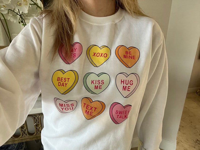 K&ouml;nnte beinhalten: Wei&szlig;es Sweatshirt mit bunten Bonbonherz-Grafiken mit Aufschriften wie "BE MINE" und "SWEET TALK". Die Herzen sind in verschiedenen Farben gehalten, darunter Rosa, Gelb, Orange und Gr&uuml;n. Das Sweatshirt hat einen Rundhalsausschnitt und lange &Auml;rmel.