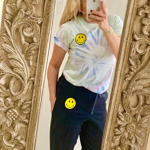 Puede incluir: Una mujer lleva una camiseta de color azul claro con estampado tie-dye y un parche con una cara sonriente amarilla. Tambi&eacute;n lleva pantalones de ch&aacute;ndal negros con un parche con una cara sonriente amarilla en la pierna izquierda.