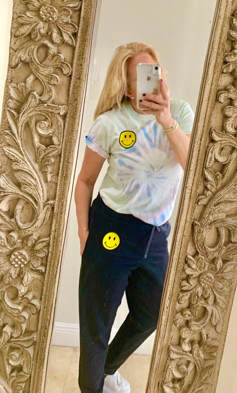 Puede incluir: Una mujer lleva una camiseta de color azul claro con estampado tie-dye y un parche con una cara sonriente amarilla. Tambi&eacute;n lleva pantalones de ch&aacute;ndal negros con un parche con una cara sonriente amarilla en la pierna izquierda.