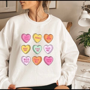K&ouml;nnte beinhalten: Wei&szlig;es Sweatshirt mit bunten Bonbonherzen, die S&auml;tze wie &bdquo;Say Yes&ldquo; und &bdquo;Kiss Me&ldquo; tragen. Die Herzen sind in verschiedenen Farben gehalten, ideal f&uuml;r den Valentinstag oder um Zuneigung auszudr&uuml;cken.