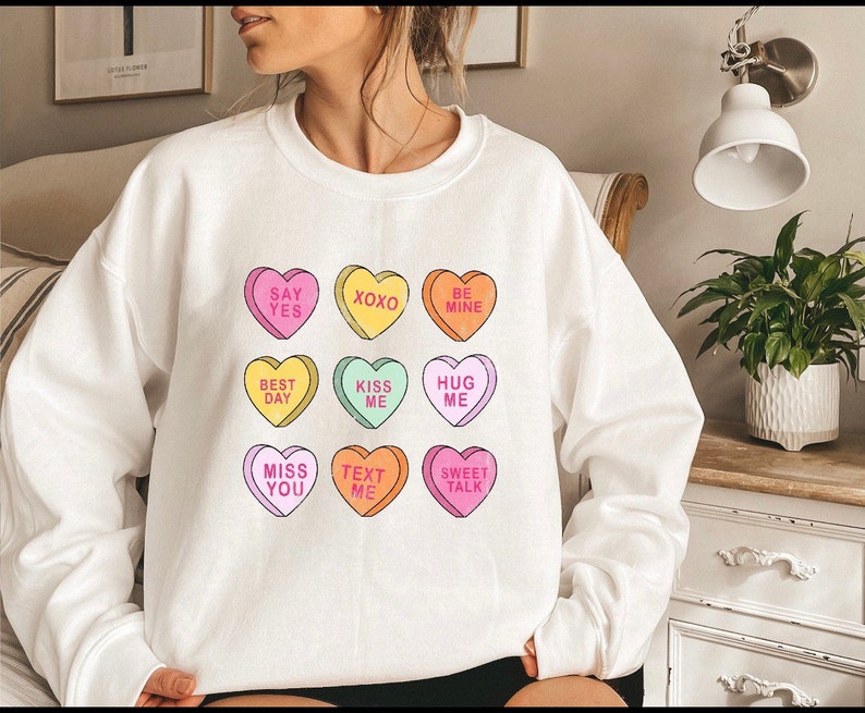 K&ouml;nnte beinhalten: Wei&szlig;es Sweatshirt mit bunten Bonbonherzen, die S&auml;tze wie &bdquo;Say Yes&ldquo; und &bdquo;Kiss Me&ldquo; tragen. Die Herzen sind in verschiedenen Farben gehalten, ideal f&uuml;r den Valentinstag oder um Zuneigung auszudr&uuml;cken.