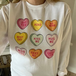 K&ouml;nnte beinhalten: Wei&szlig;es Langarm-Sweatshirt mit neun bunten Bonbonherzen. Jedes Herz tr&auml;gt eine andere Botschaft, darunter "Say Yes", "XOXO" und "Be Mine". Ein verspieltes Design, ideal f&uuml;r den Valentinstag oder um Zuneigung auszudr&uuml;cken.