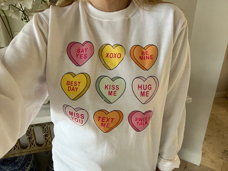 K&ouml;nnte beinhalten: Wei&szlig;es Langarm-Sweatshirt mit neun bunten Bonbonherzen. Jedes Herz tr&auml;gt eine andere Botschaft, darunter "Say Yes", "XOXO" und "Be Mine". Ein verspieltes Design, ideal f&uuml;r den Valentinstag oder um Zuneigung auszudr&uuml;cken.