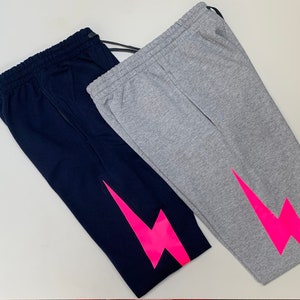 Puede incluir: Dos pantalones de ch&aacute;ndal con un dise&ntilde;o de rayo rosa. Un pantal&oacute;n es azul marino y el otro es gris.