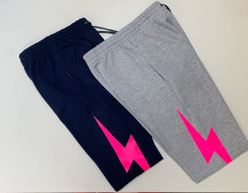 Puede incluir: Dos pantalones de ch&aacute;ndal con un dise&ntilde;o de rayo rosa. Un pantal&oacute;n es azul marino y el otro es gris.